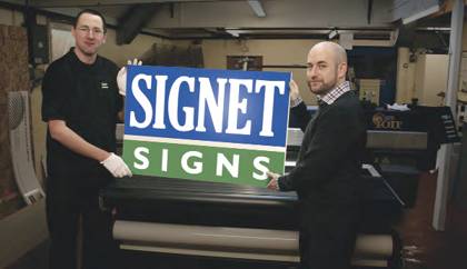 2 people holding up a Signet Signs sign.