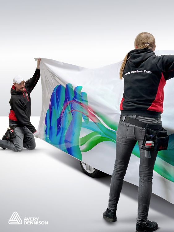 2 people holding up a sheet of SP 1504 self-adhesive film and laying it onto a car for wrapping