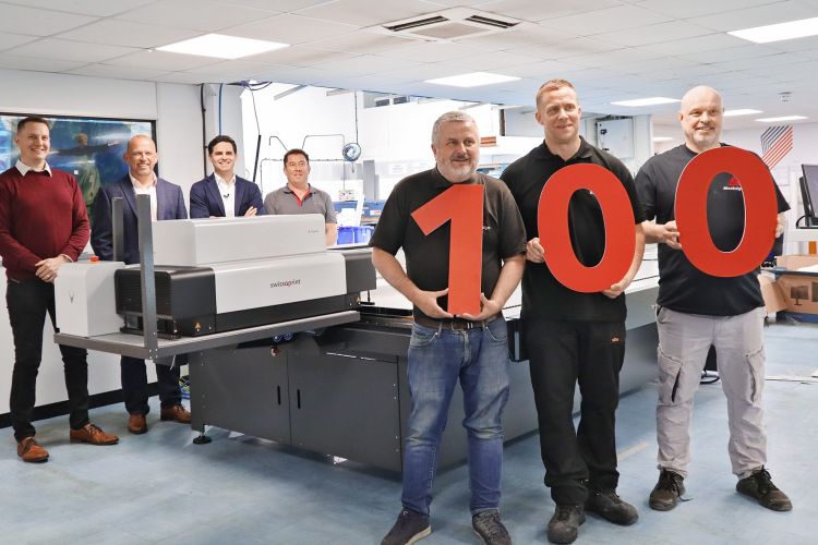 Three men stand at the front of the image holding the number 100. Behind them is the Impala 4 flatbed printer and the staff members at Stocksigns