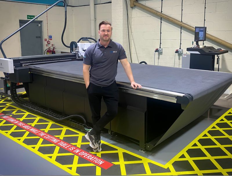A man leans with a hand in his pocket, smiling against the new purchase of the Kongsberg digital cutting table