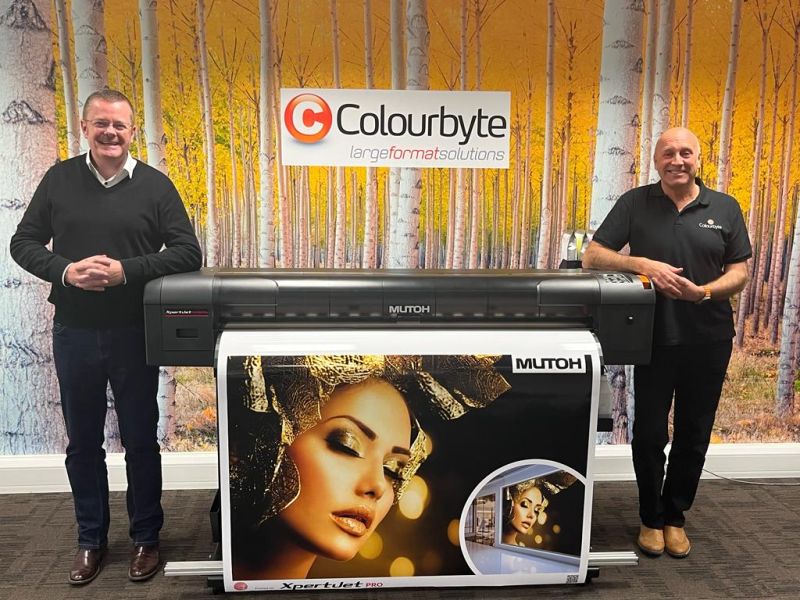 Two men stand either side of a printer with Colourbyte logo against the wall behind them