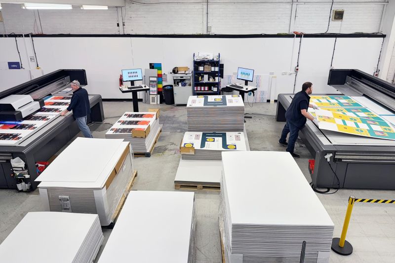 A large workshop with two flatbed printers facing each other and stock items piled high next to them, ready for print