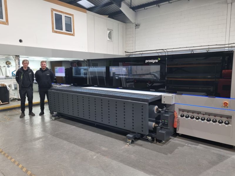Two men stand next to a wide format printer as it is printing onto media